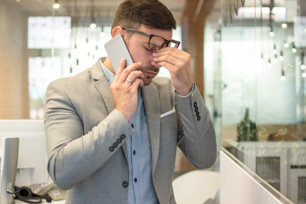 A professional man showing signs of stress with a phone on one hand, closing his eyes and his other hand holding his nose in between his eyes.